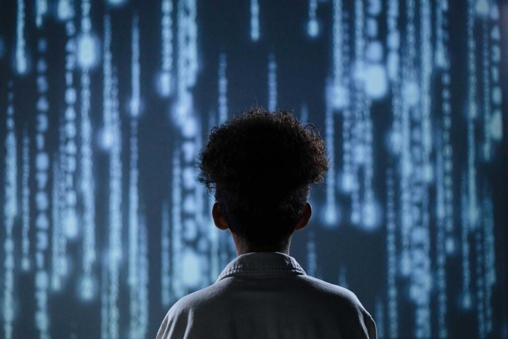 Photo of person of colour in silhouette with back facing camera, staring at cascade of computer data similar to the Matrix.