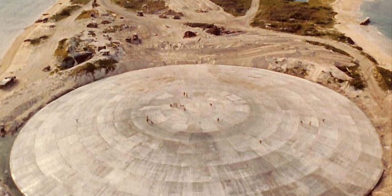 Runit Dome sits roughly 25 feet above sea level on low-lying Runit Island, making it vulnerable to inundation from rising seas. People on top of the thick concrete cap, which measures nearly 400 feet in diameter, look like ants in this aerial view. (Photo from Wikimedia Commons)